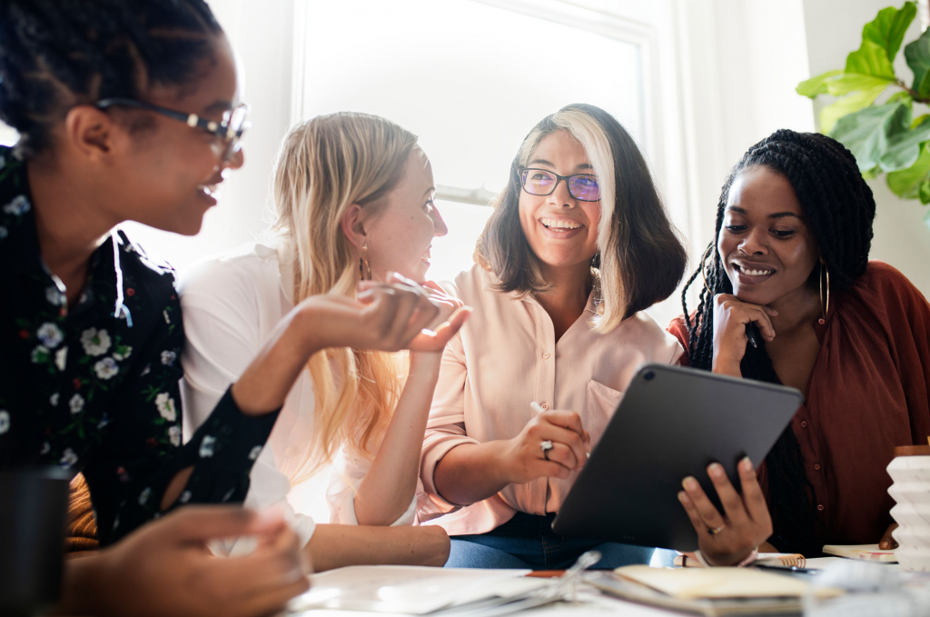 Group of four seated women facing each other. One woman is holding a tablet device in her hand.