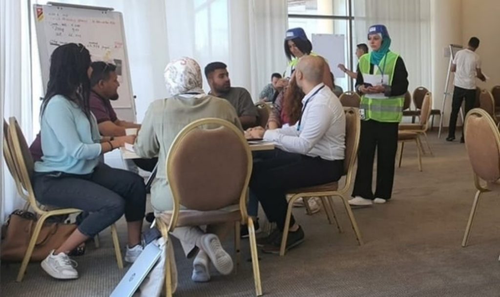 Group of five men and women are sitting on the chairs around the table, listening to two women in light reflecting vests