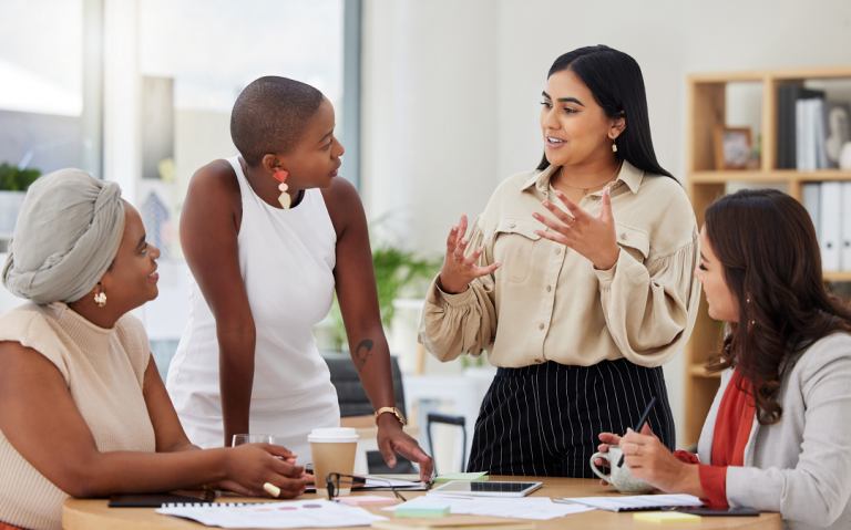 The featured image shows a group of four women in an office setting. Two are seated and two are standing. One of the standing women is talking while the others face her.